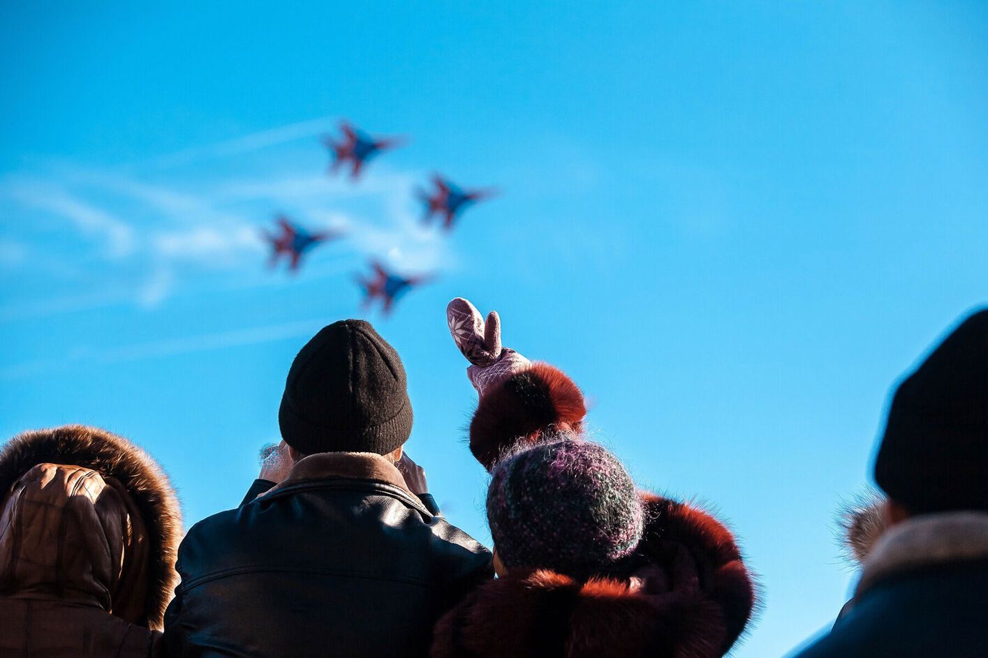 People watching jets fly in formation against a blue sky, one person waves.