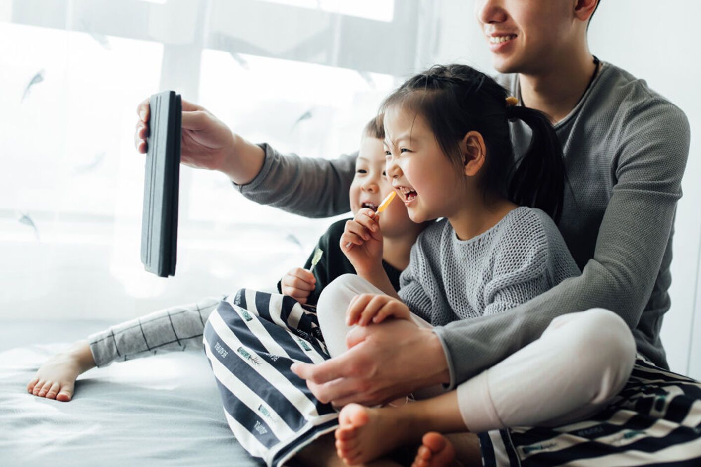 Father and two children looking at a tablet. They sit on a bed near a window, smiling.