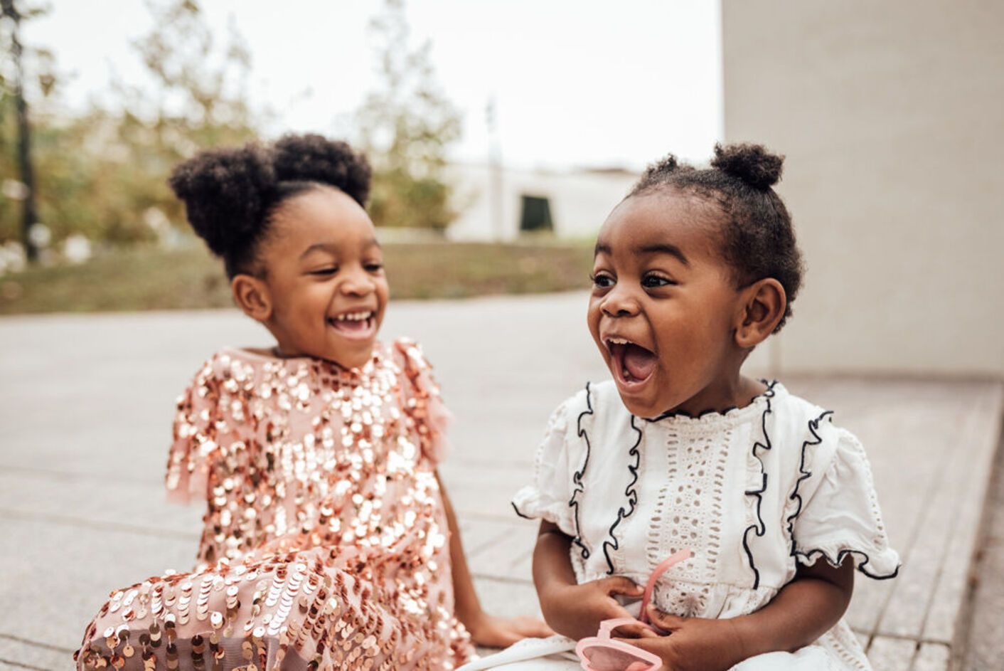 Two Black children laughing, one in a sparkly pink dress, the other in a white dress, outdoors.