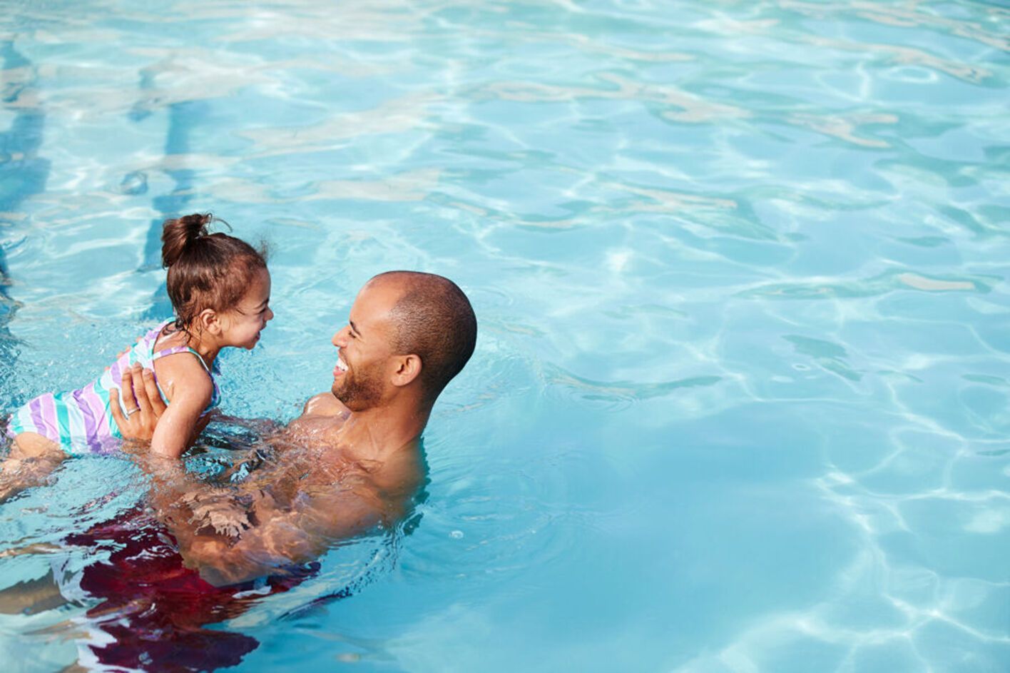 Father and daughter in a pool, smiling. Father holds the child, both in swimwear, blue water.