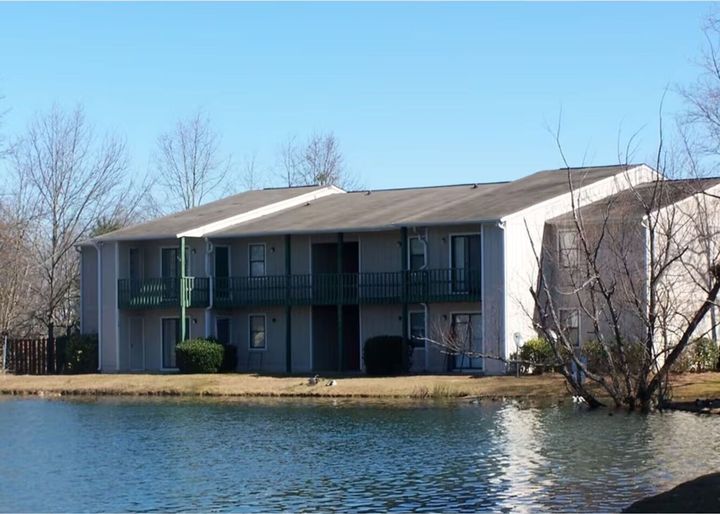 Two-story gray apartment building with balconies on the edge of a lake, under a blue sky.