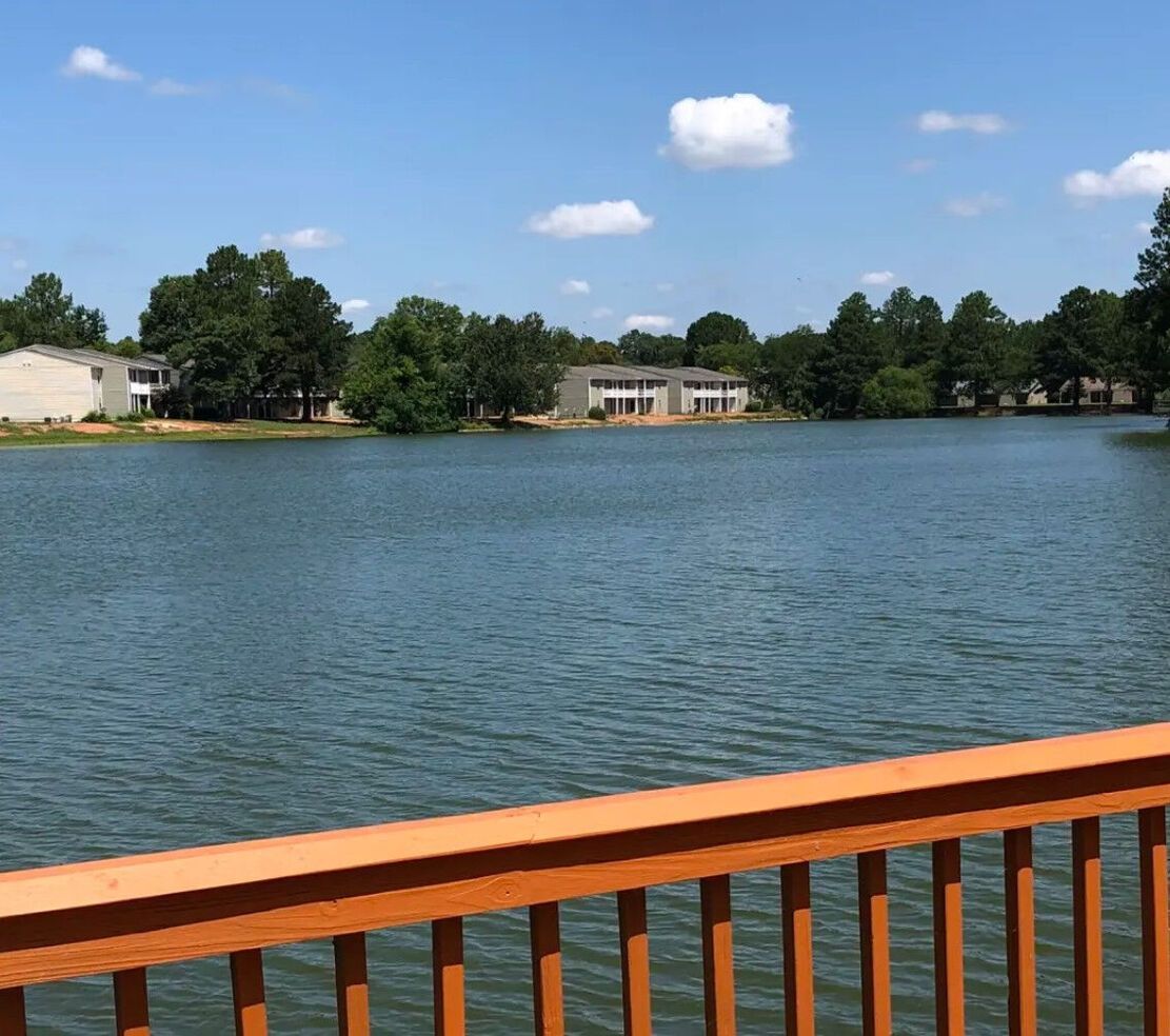A lake view with homes, trees, and a wooden railing on a sunny day.