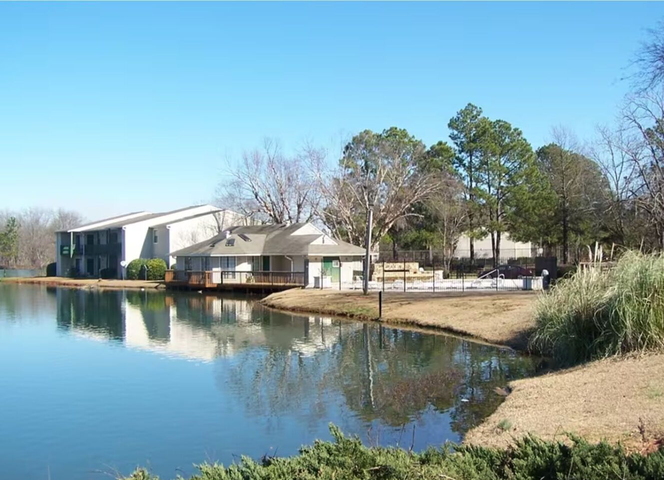 Lakeside buildings with trees and blue sky reflected in calm water.