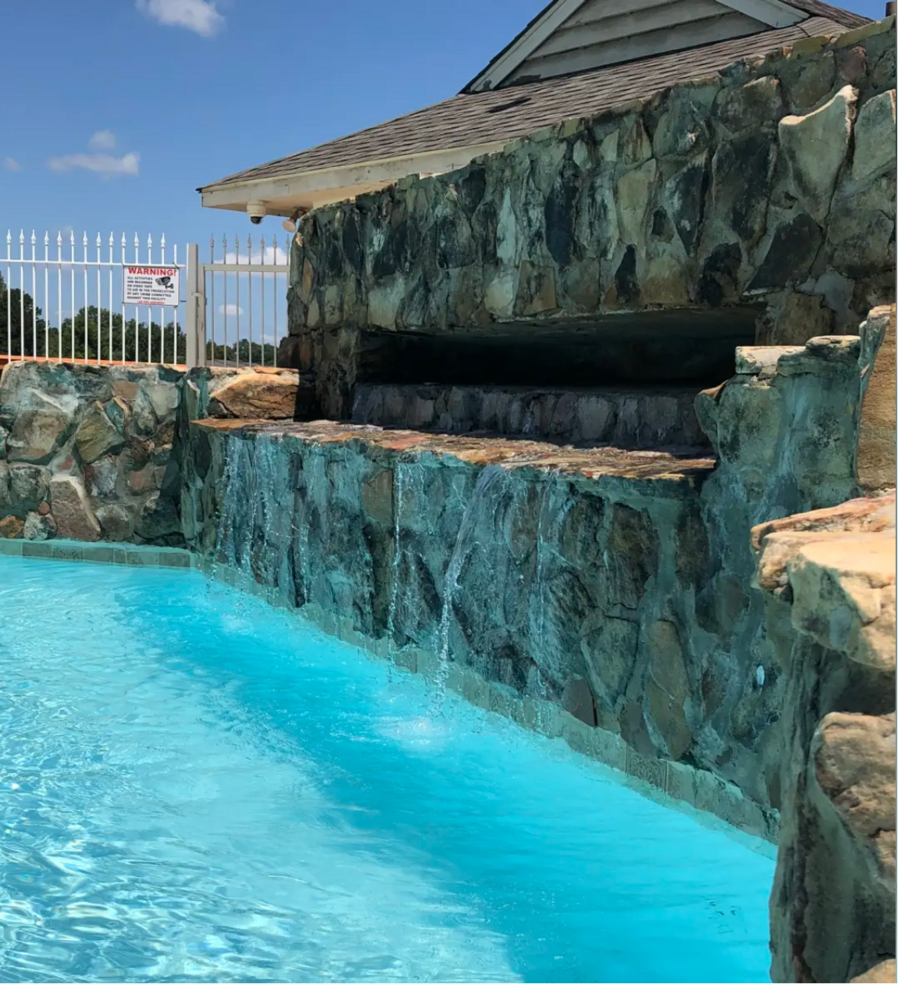 Waterfall feature cascading into a bright blue swimming pool. Stone facade, white fence, blue sky.