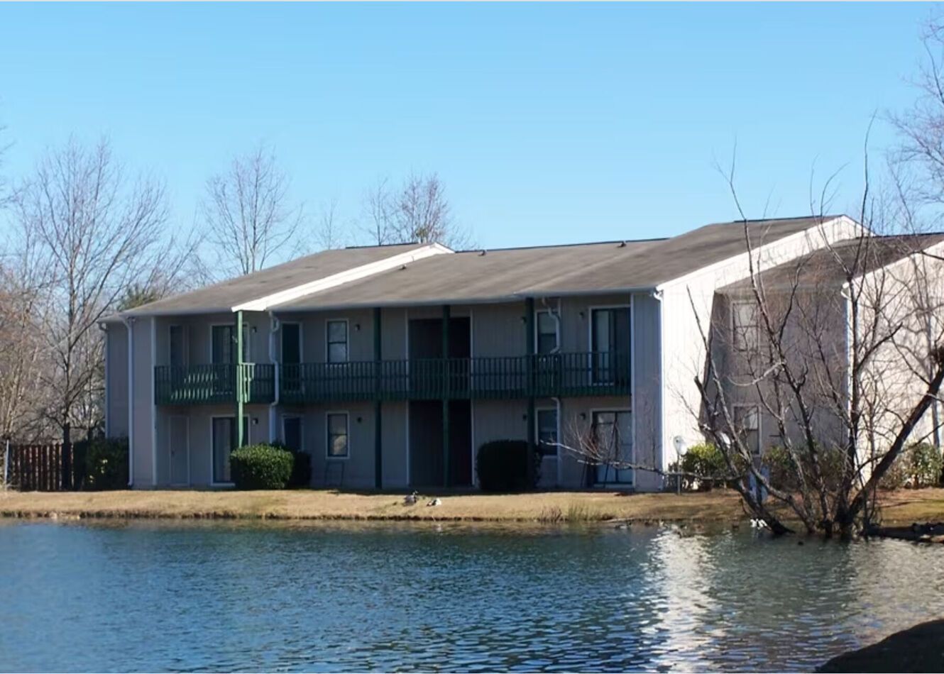 Two-story apartment building on the edge of a pond, blue water, green balconies, sunny day.