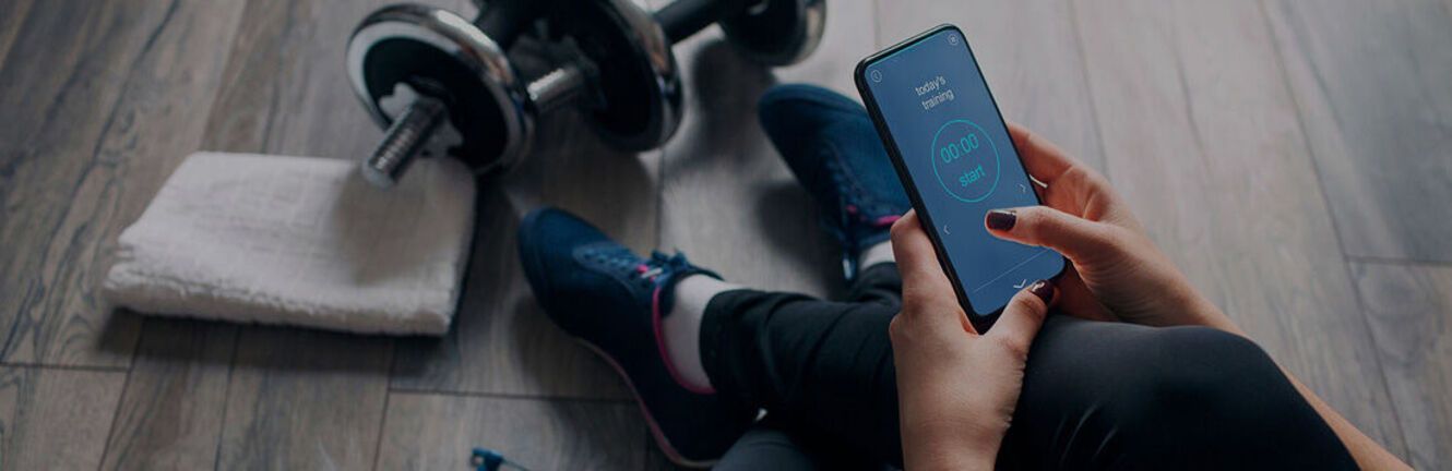 Person sits with phone, weights, and a towel on wooden floor.