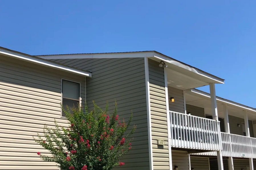 Apartment building with tan siding, white trim, and a balcony under a blue sky.