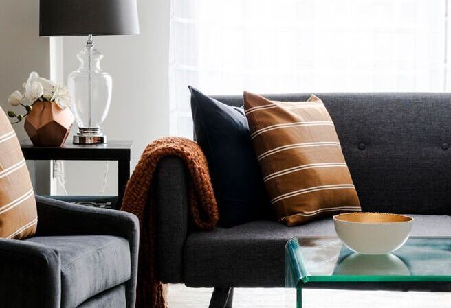 Living room with gray sofas, throw pillows, a blanket, a table lamp, and a glass coffee table.
