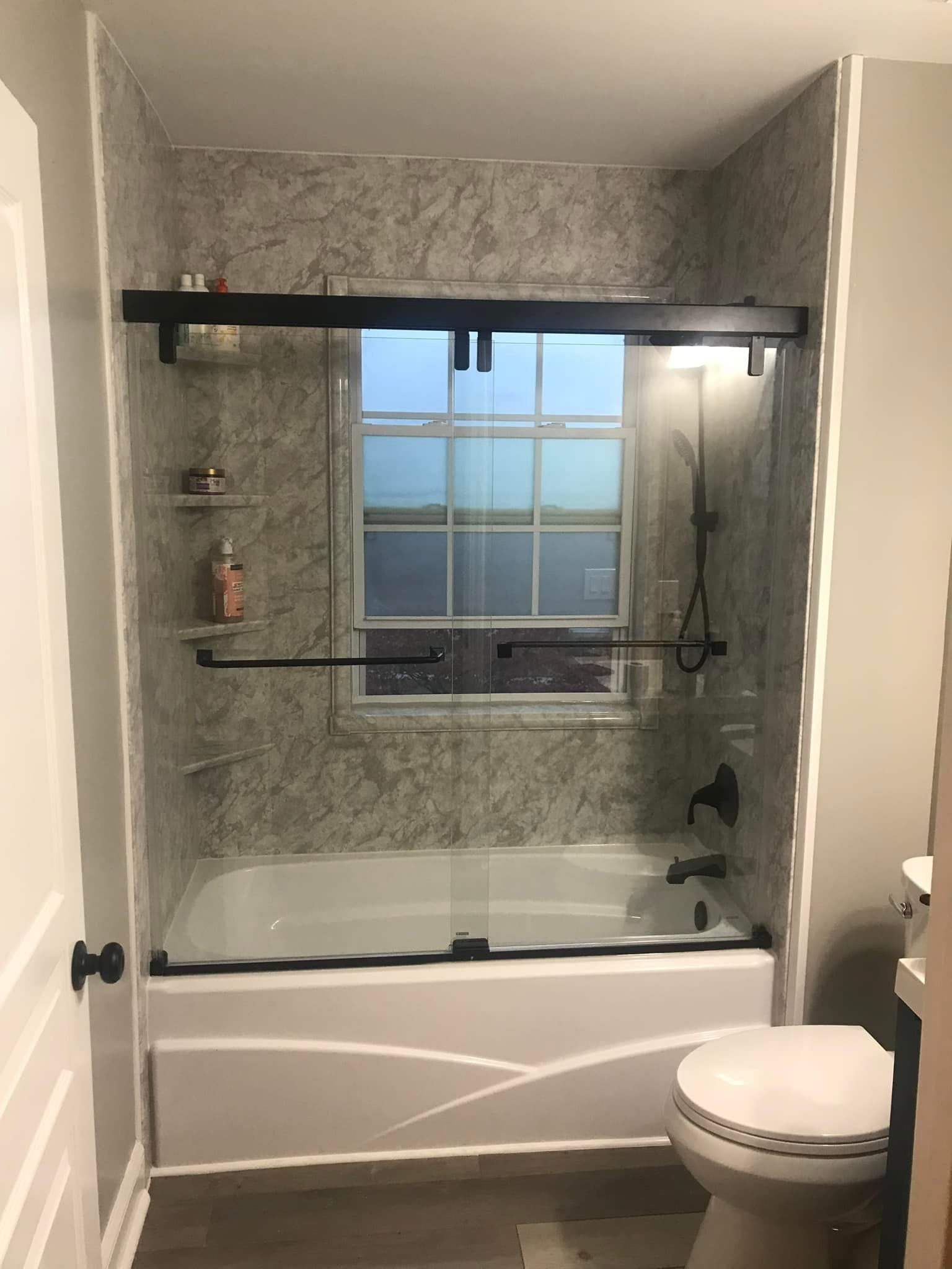 Bathroom with a tub, shower doors, and a window. Gray tile and black fixtures.