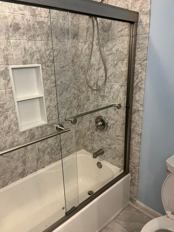 Bathroom with gray tile, shower with glass door, tub, and grab bars.
