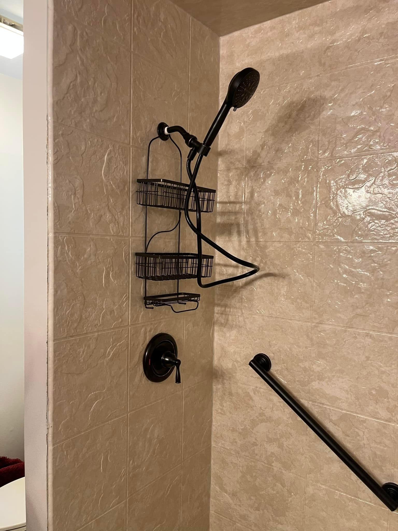 Shower stall with bronze showerhead, caddy, and grab bar on textured beige tile.