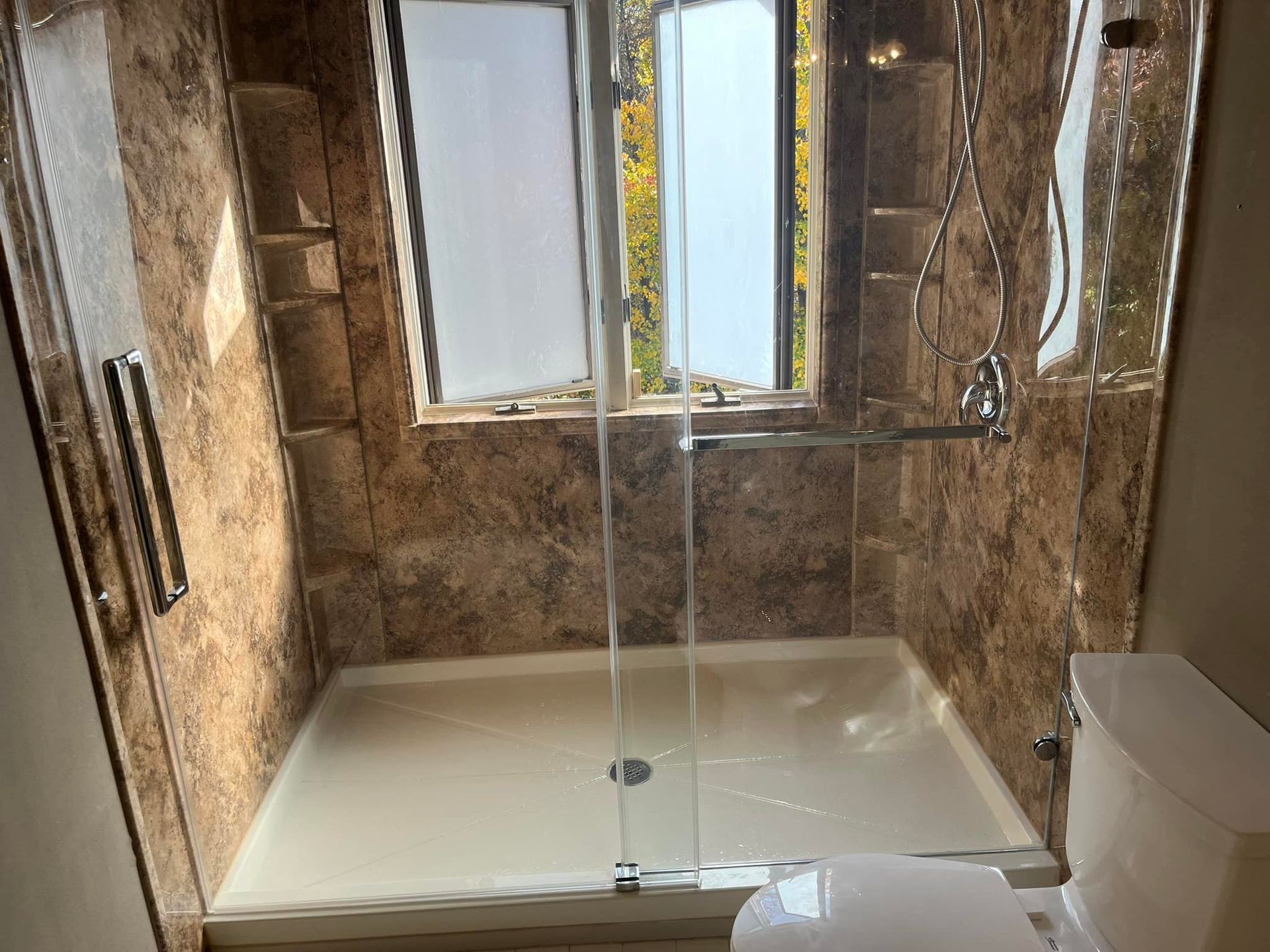 Shower with brown marbled walls, glass door, window, and toilet.