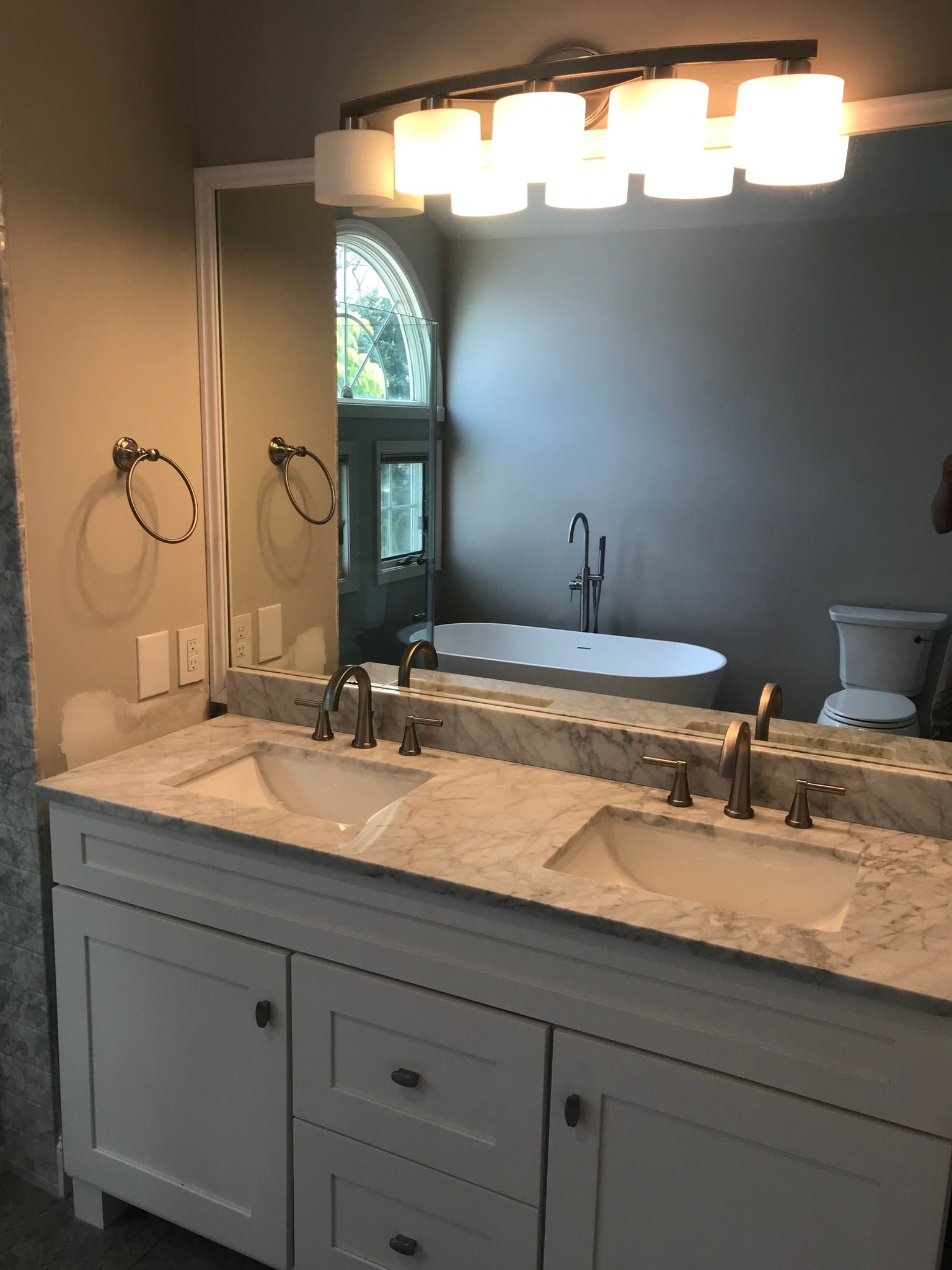 Bathroom with white vanity, double sinks, large mirror, and a tub in the background.