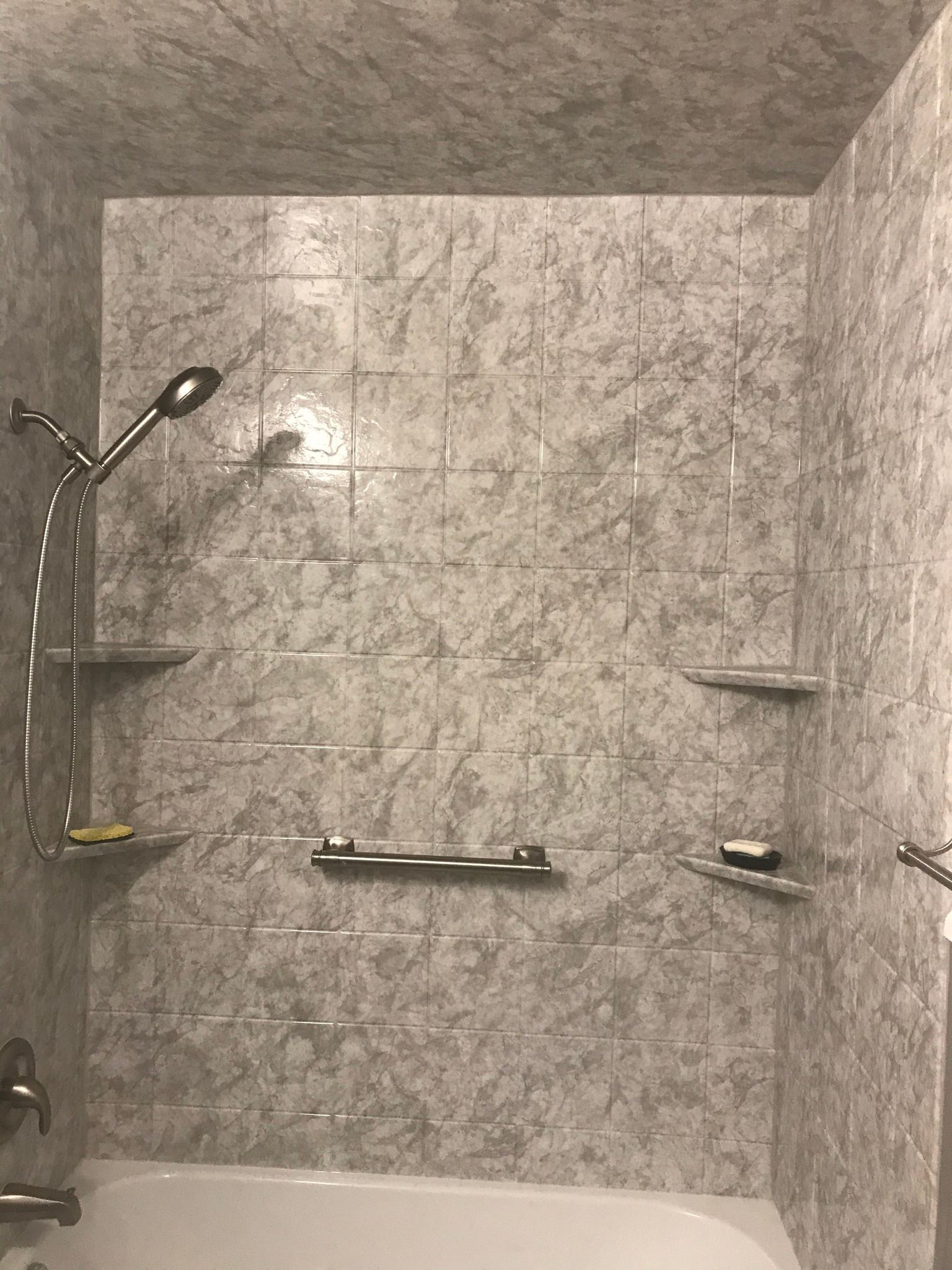 Gray tiled shower with a showerhead, shelves, and a grab bar above a white bathtub.