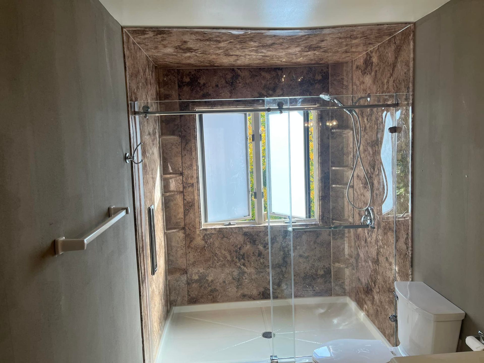 Bathroom with a marble-look shower, glass doors, a toilet, and a window behind the shower.