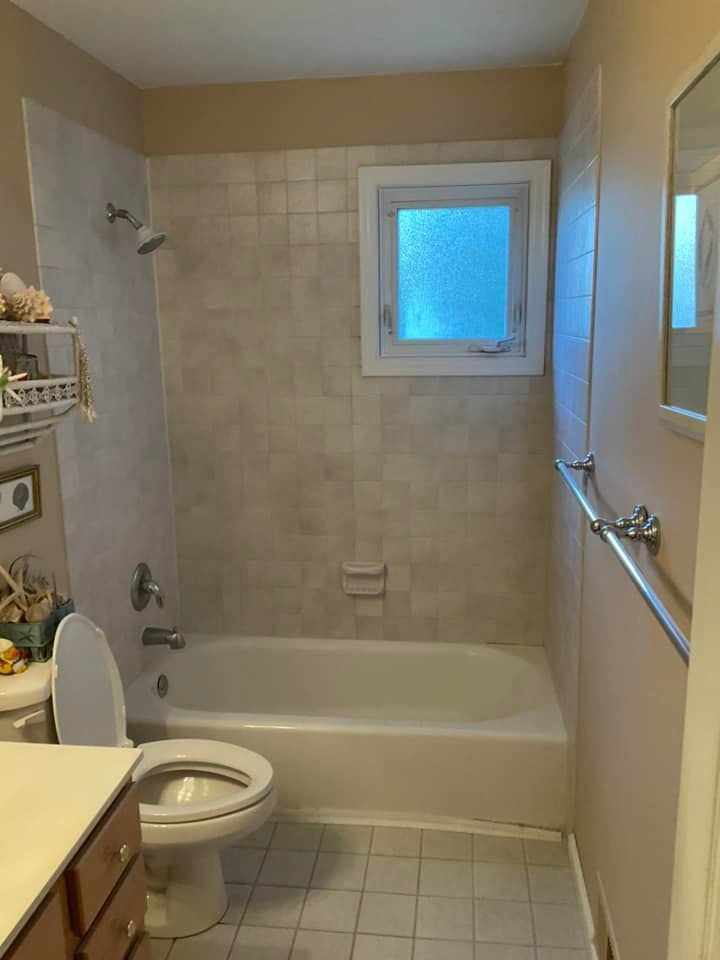 Bathroom with a tub, toilet, window, and handrail. Beige walls and tile.