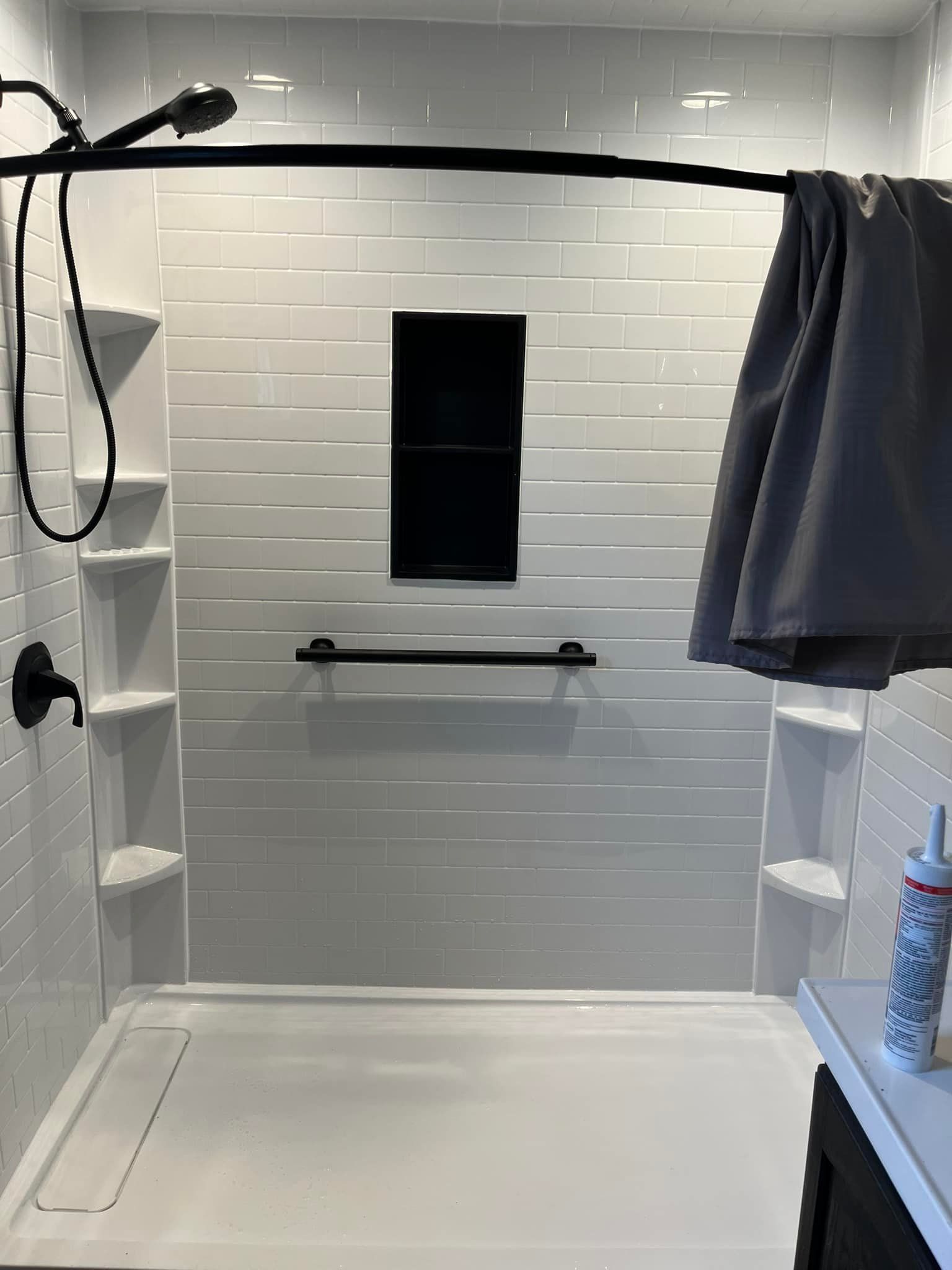 White tiled shower with built-in shelves, black fixtures, and a gray towel, with a dark cabinet visible.