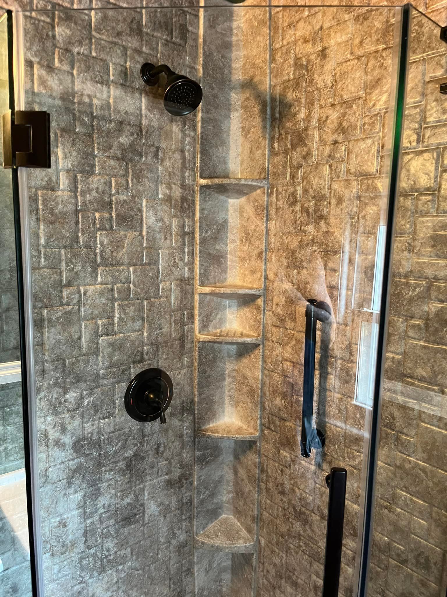 Shower with textured gray walls, dark fixtures, glass door, and built-in shelves.