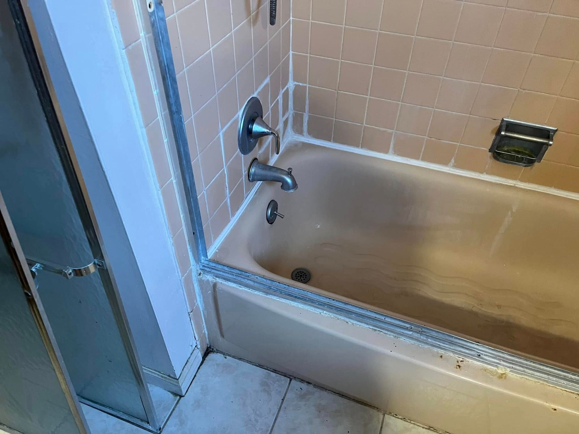 Beige bathtub with tan tiles, chrome fixtures, and glass shower door.