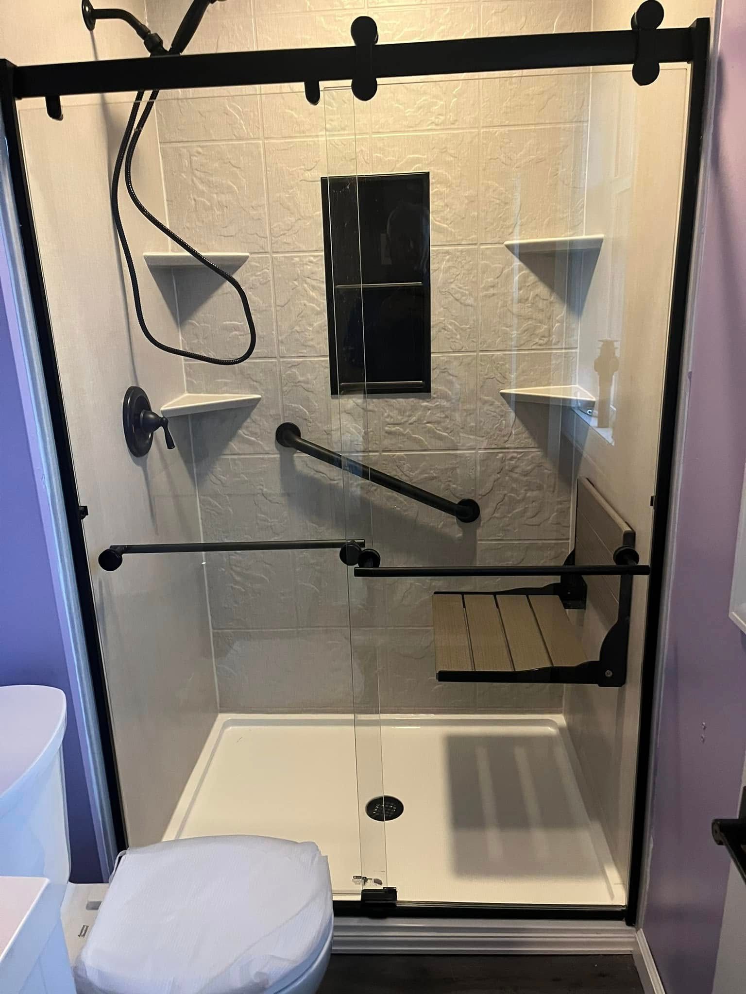 Accessible shower with grab bars, seat, black frame, and white and gray tiles.