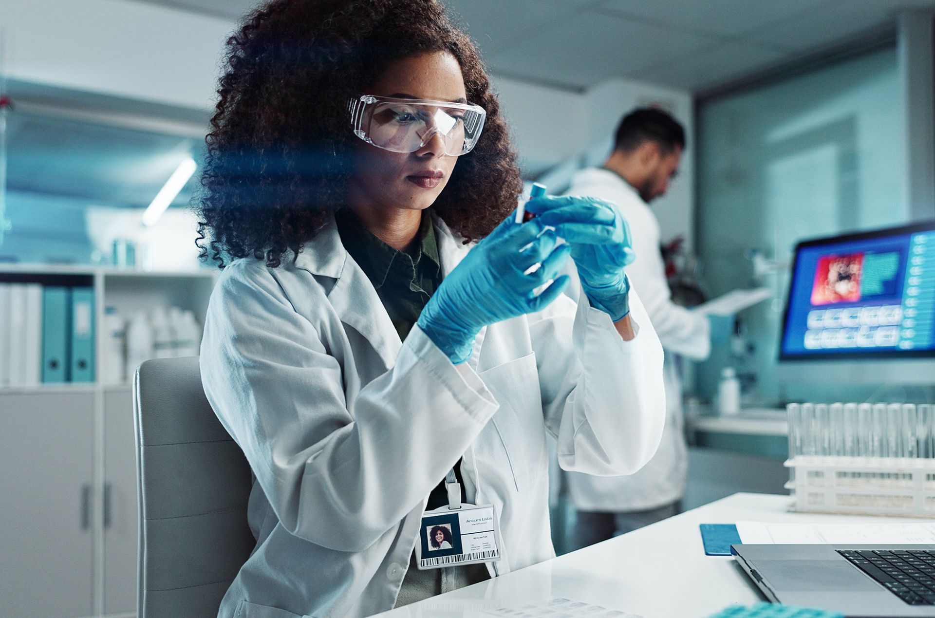 A woman in a lab coat and gloves is holding a test tube in a laboratory.