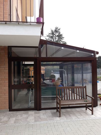 Patio chiuso con pareti in vetro e una panca in legno all'esterno, collegato a un edificio con balcone.