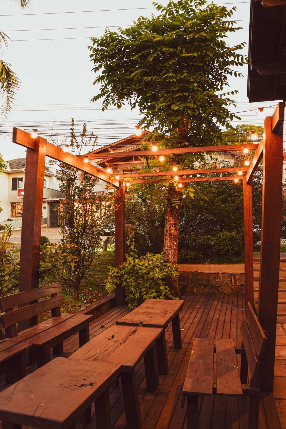 Um deck de madeira com mesas e bancos sob uma pérgola com luzes.