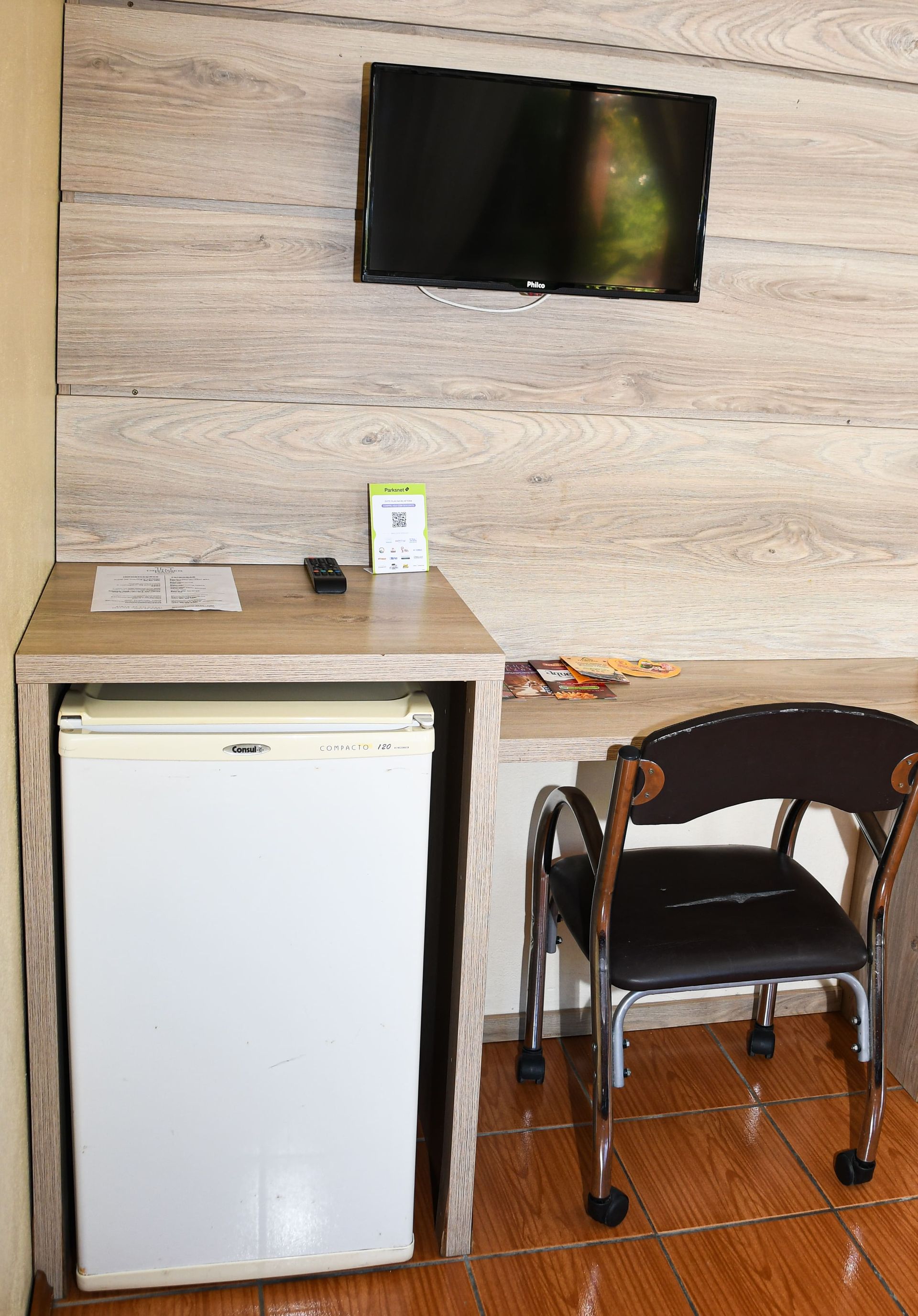 Uma sala com escrivaninha, cadeira, geladeira e televisão.