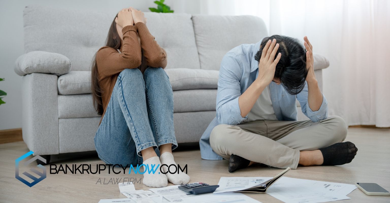Couple sits on floor, stressed, hands on faces near bills and calculator.