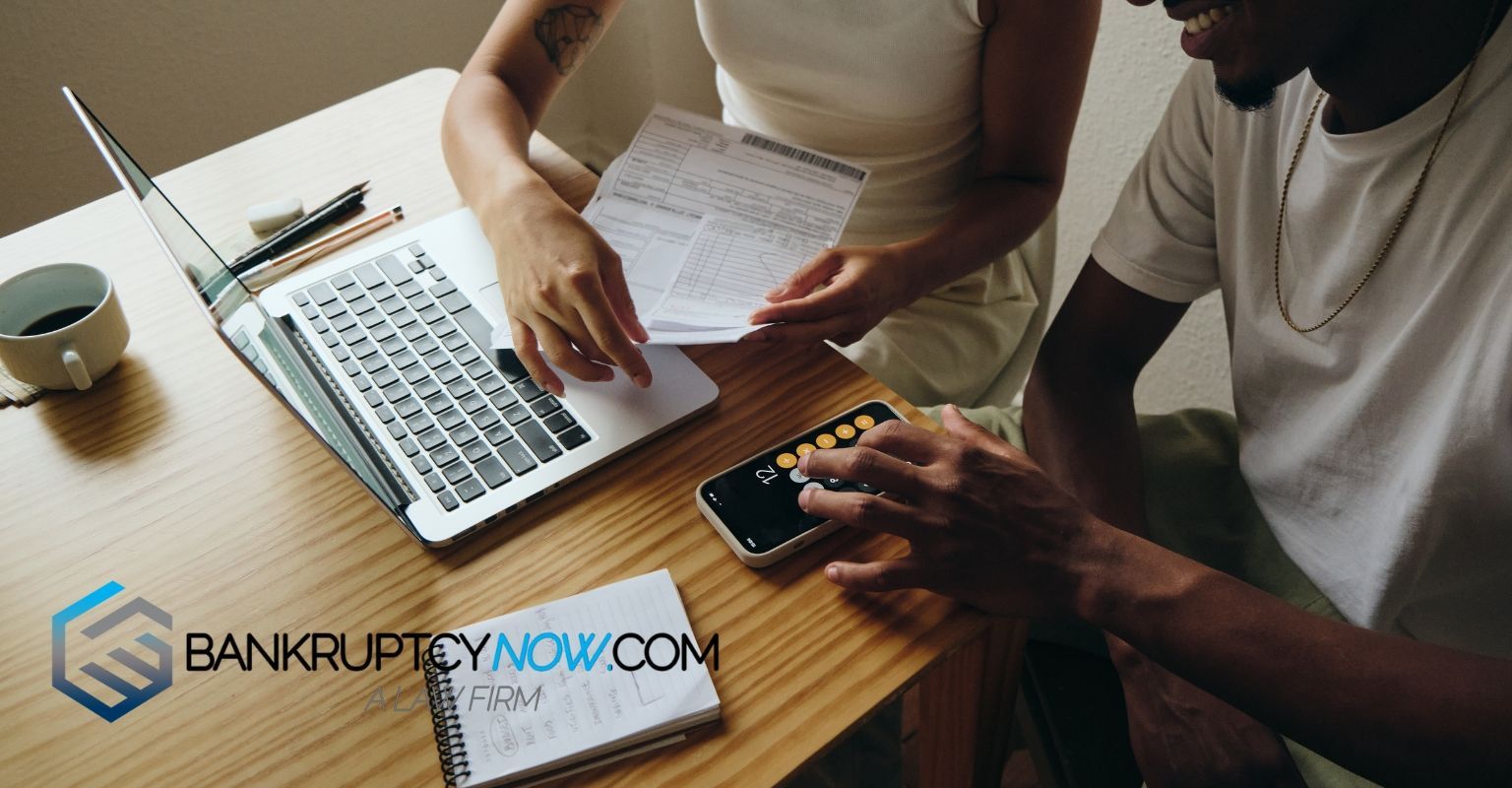 Couple reviews documents at table with laptop, calculator, and notebook; logo: BankruptcyNow.com.