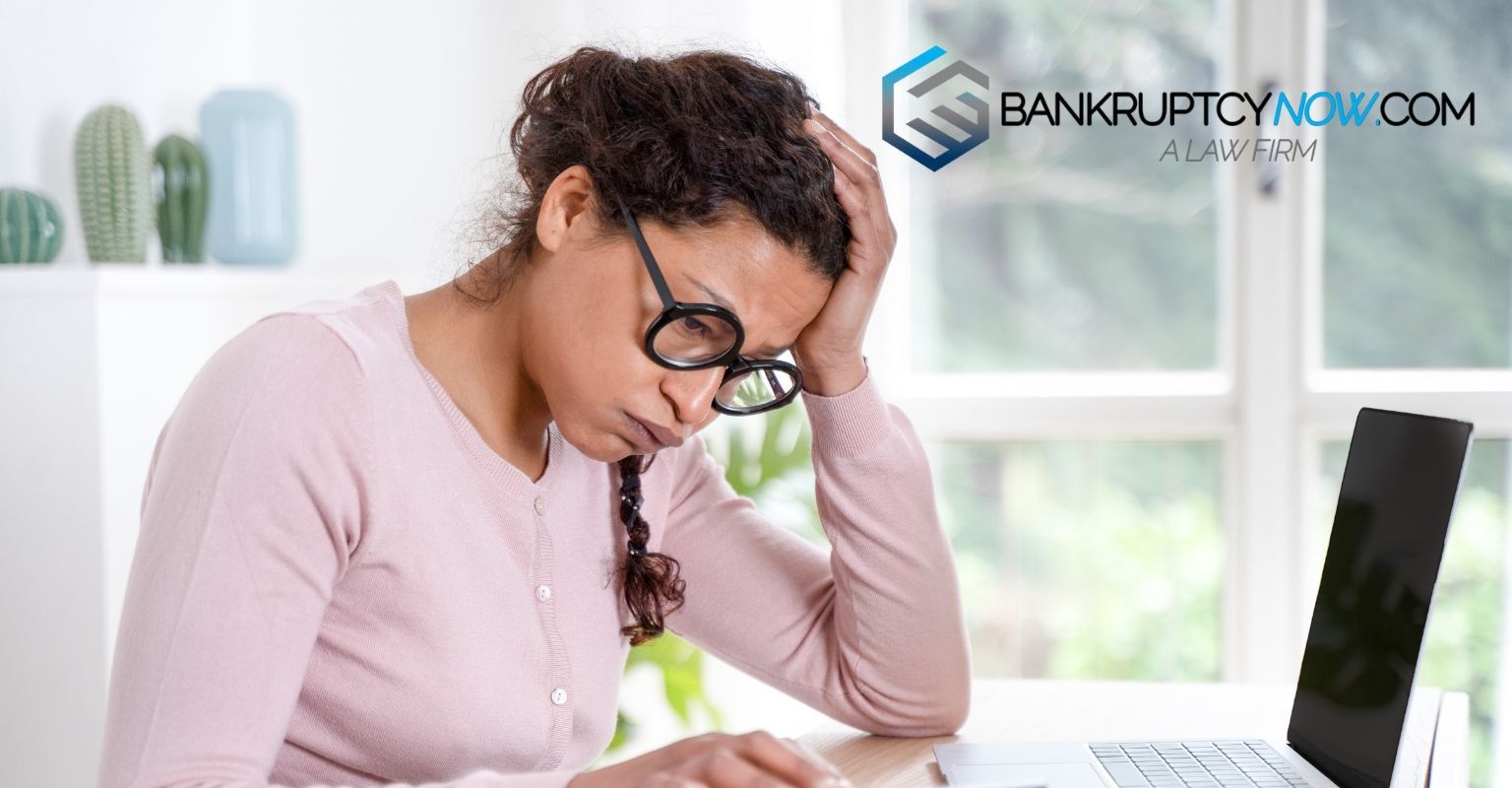 Woman with glasses looking stressed, holding head, sitting at a laptop. Logo 