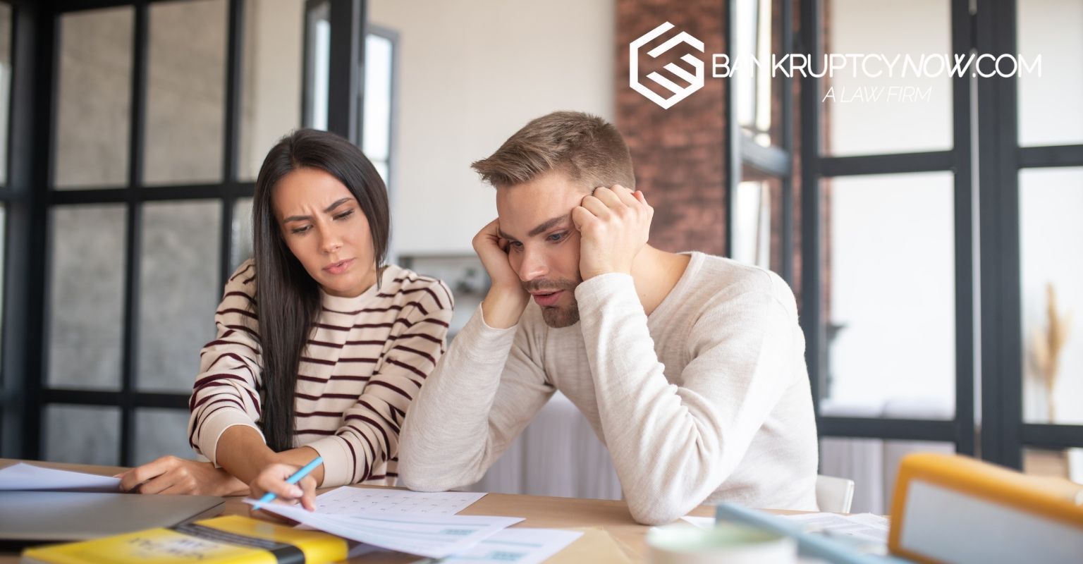A couple looking at papers, appearing stressed. Man holding his head, woman pointing. Bright room.