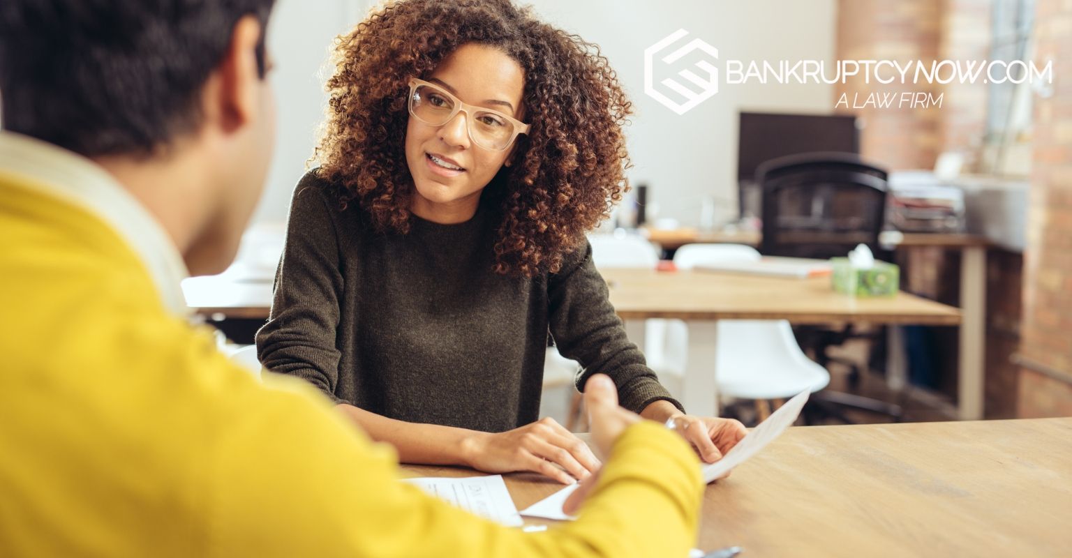 Woman in glasses talking to person holding papers at a desk, office setting.