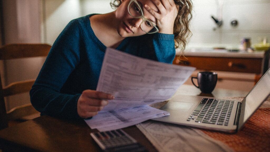 Woman with glasses looks stressed, holding paperwork, laptop and calculator nearby.