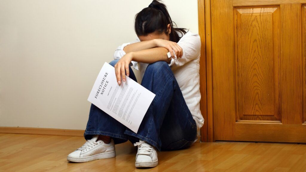Woman sitting on floor, head in arms, holding a test paper, near a door.