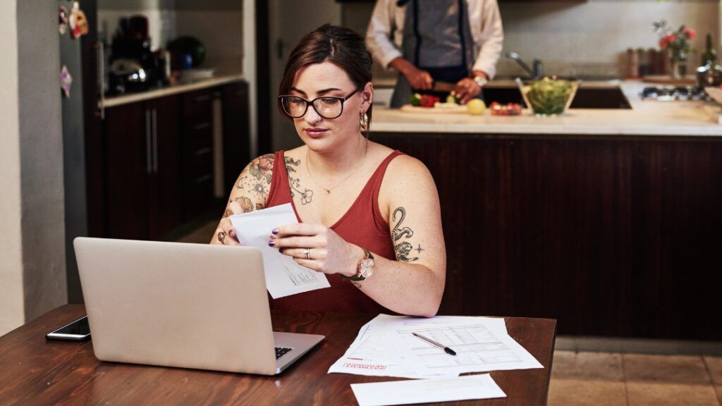 Woman at table with laptop reviews bills, man cooks in background.