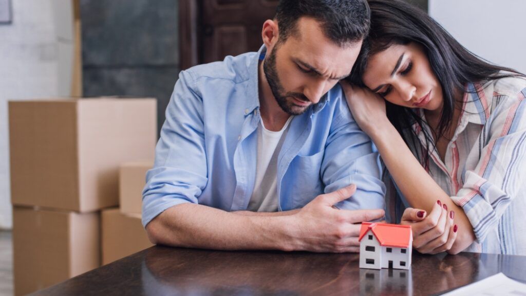 Couple sadly looks at a small house model, cardboard boxes in the background.