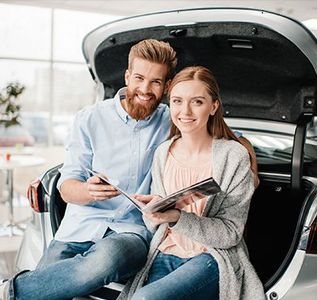 Couple seated in car trunk, smiling, holding brochure in showroom.