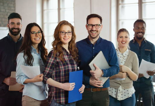 Group of six diverse coworkers smiling at the camera in an office setting, some holding papers and a laptop.