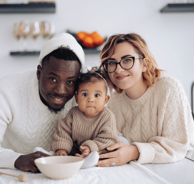 A family of three, smiling at the camera, sitting at a table. Includes a Black man, a White woman with glasses, and a baby.
