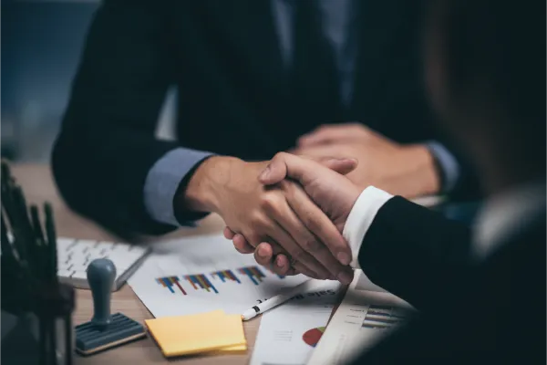 Two businesspeople in suits shaking hands over a desk with papers and a stamp.