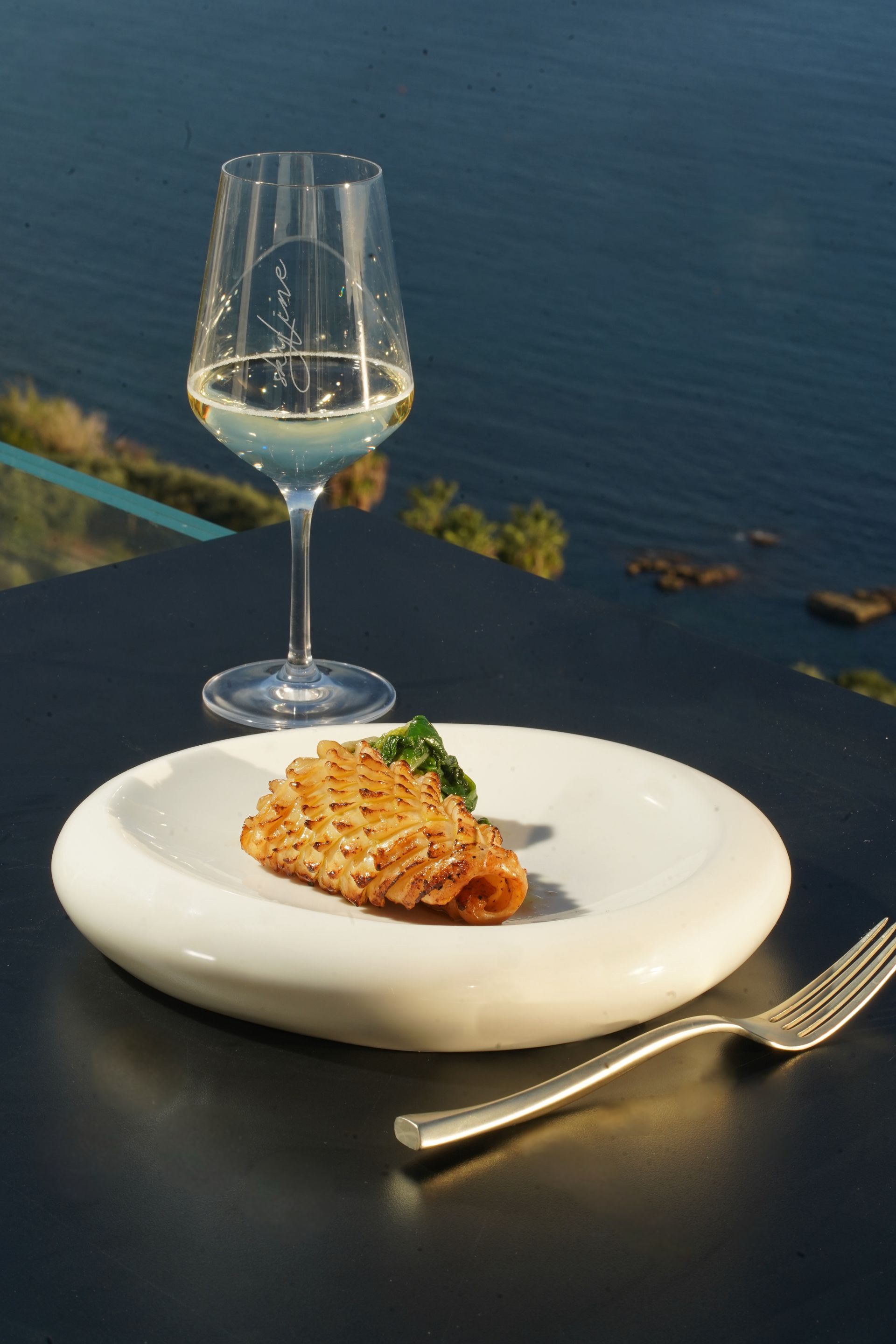 Un piatto di pasta blu con un bicchiere di vino bianco su un tavolo con vista sulla baia al tramonto.