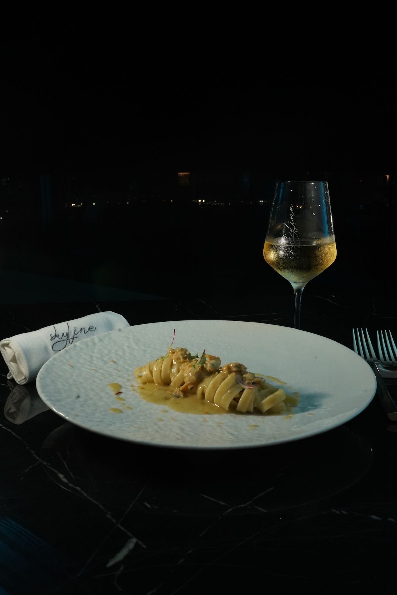 Un piatto bianco con pasta, un bicchiere di vino bianco e un tovagliolo arrotolato su un tavolo buio di notte.
