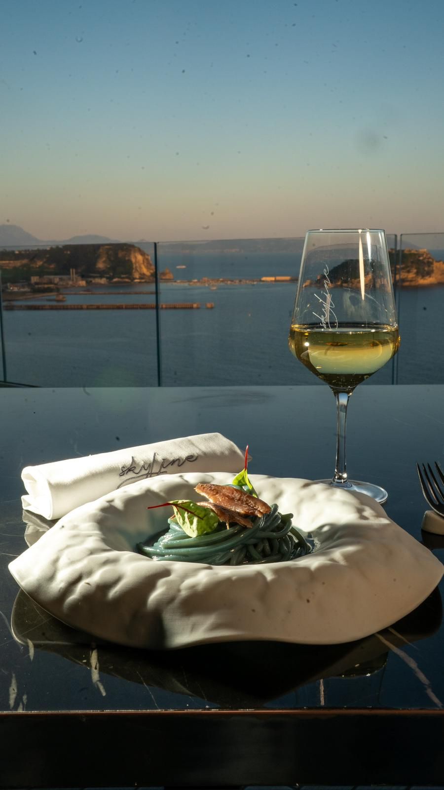 Un piatto di pasta blu con un bicchiere di vino bianco su un tavolo con vista sulla baia al tramonto.