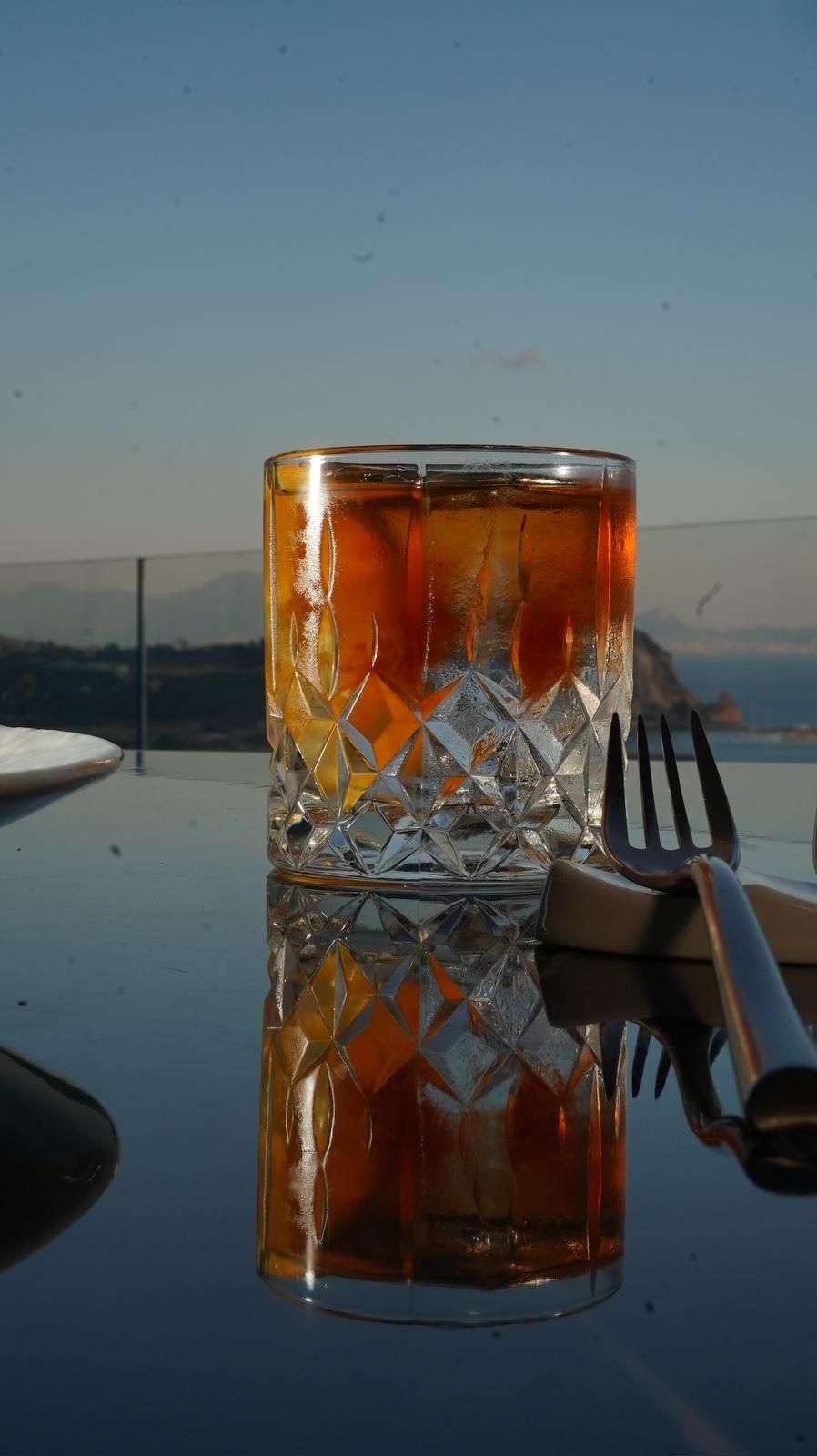 Cocktail vecchio stile con scorza d'arancia in un bicchiere tagliato su un tavolo riflettente, con l'oceano sullo sfondo.