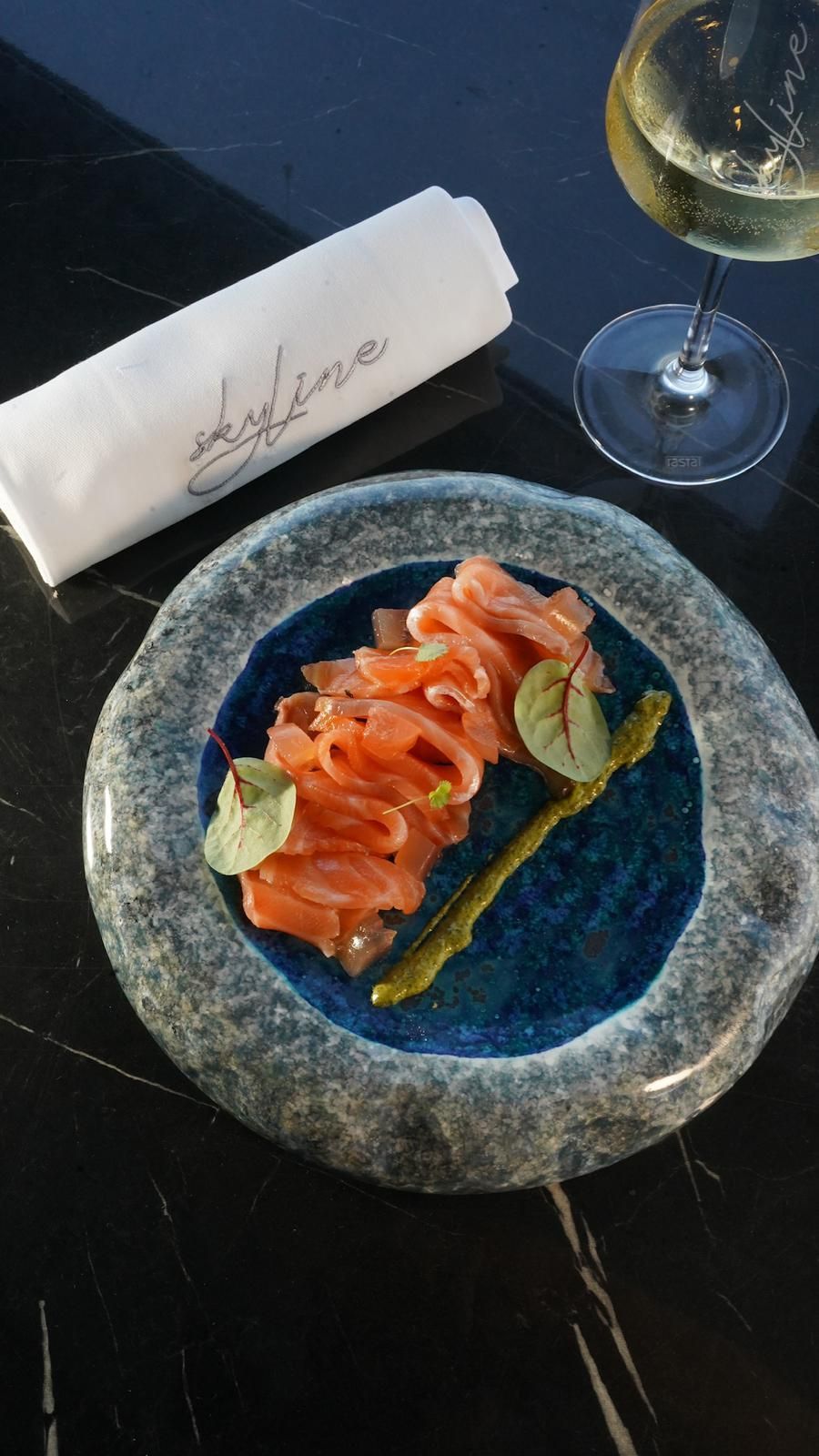 Antipasto di salmone su un piatto di pietra con bordo blu, con un tovagliolo piegato e un bicchiere di vino su un tavolo nero.