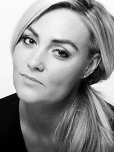 A black-and-white close-up portrait of a person with blonde hair pulled back, looking directly at the camera.