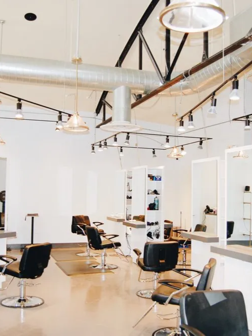 A bright, modern hair salon featuring multiple black styling chairs, mirrors, and industrial ceiling lights and ductwork.