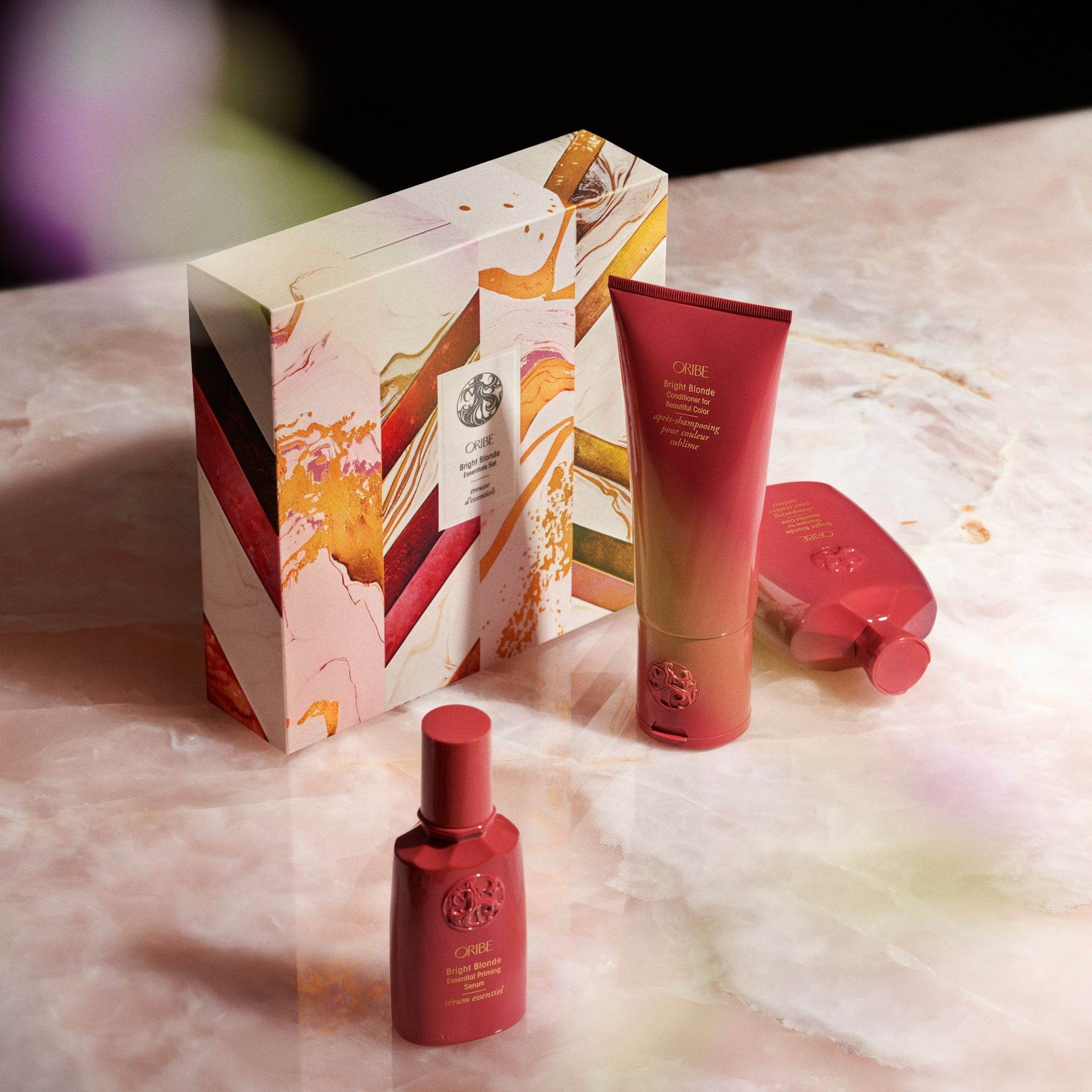 A gift set featuring a box, a tube, and a bottle of beauty products in shades of pink, orange, and red on a marble base.