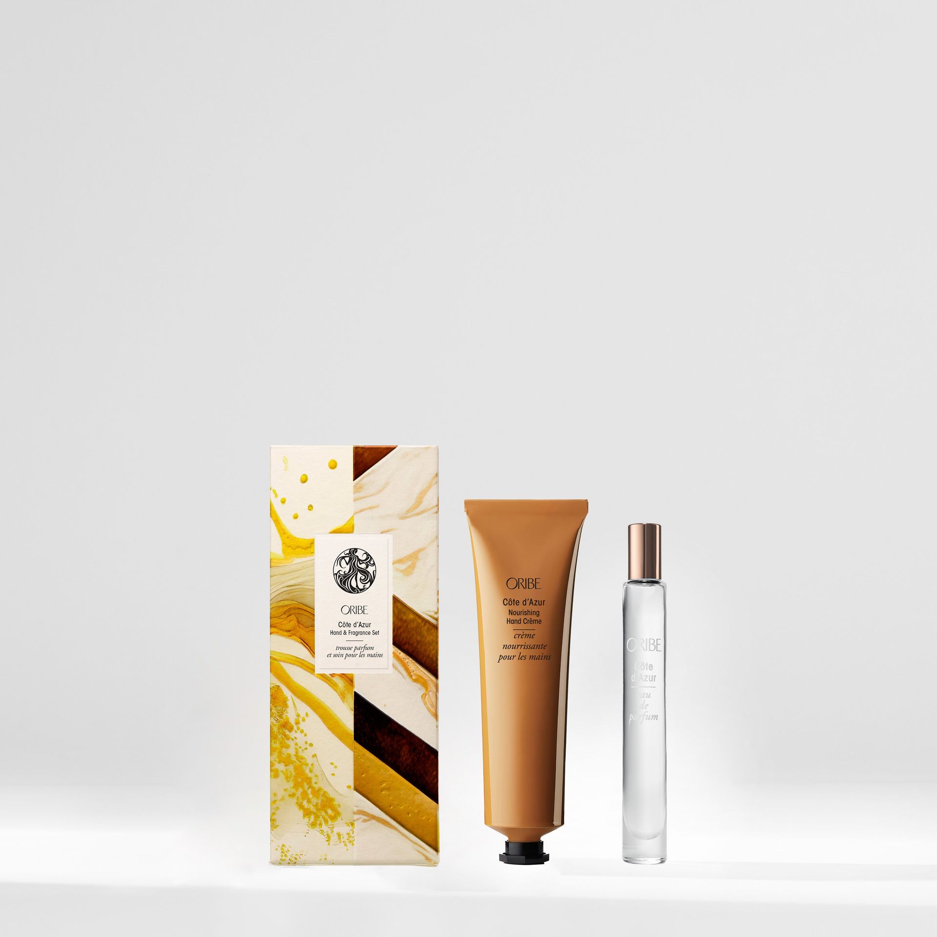 A beauty gift set featuring a patterned box, a tan tube of Oribe hand cream, and a tall, clear glass perfume bottle.